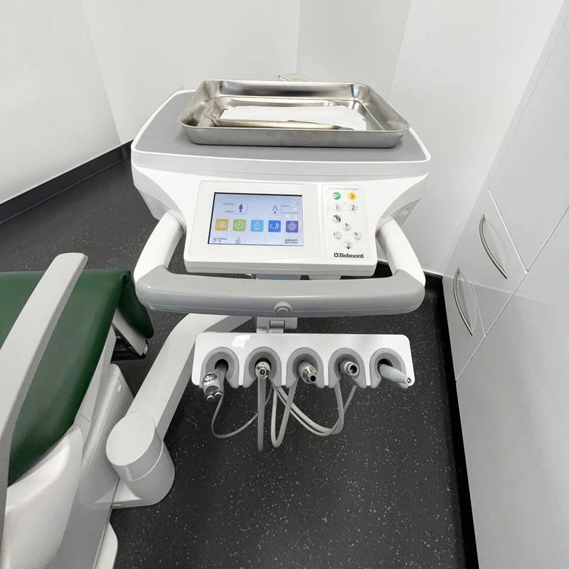 A Belmont dental operatory treatment station with a digital control screen, multiple instrument ports, and a stainless steel tray on top, shown next to a green-and-white dental chair in a clinical room.