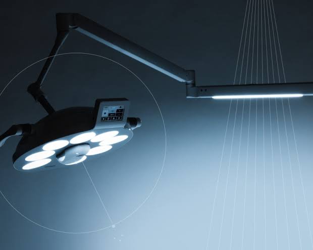 Modern surgical operating room light fixture with multiple circular bulbs and an articulated arm, shown against a dark blue-gray background with light beams and overlay graphics.